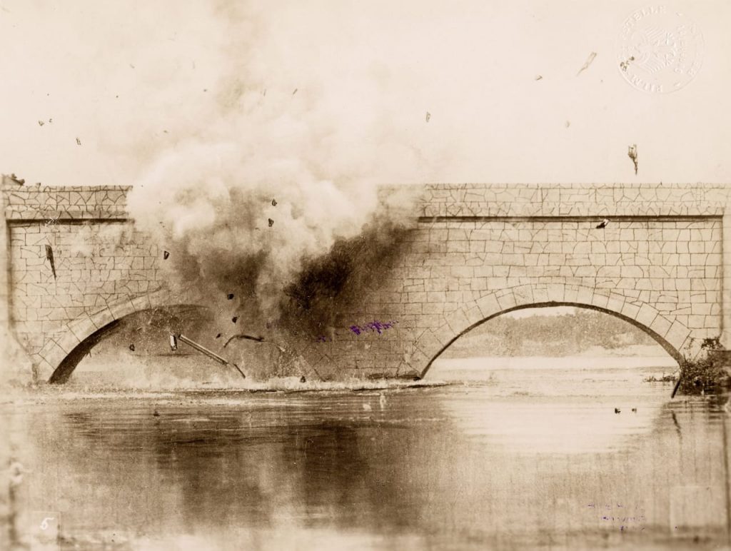 Eine Brücke überspannt einen Fluss, in Fahrbahnhöhe: eine Rauchwolke und Trümmerteile einer Explosion.