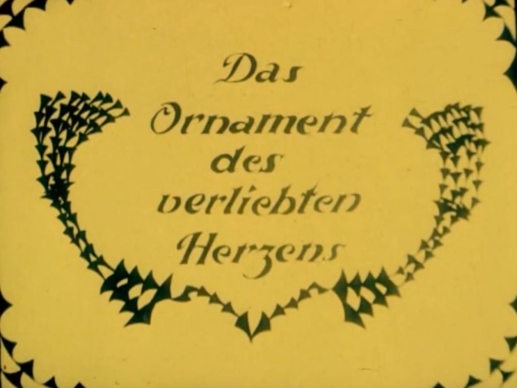 Filmstill aus Lotte Reinigers Erstlingswerk. Scherenschnitt-Ornamente umranken den Titel "Das Ornament des verliebten Herzens"